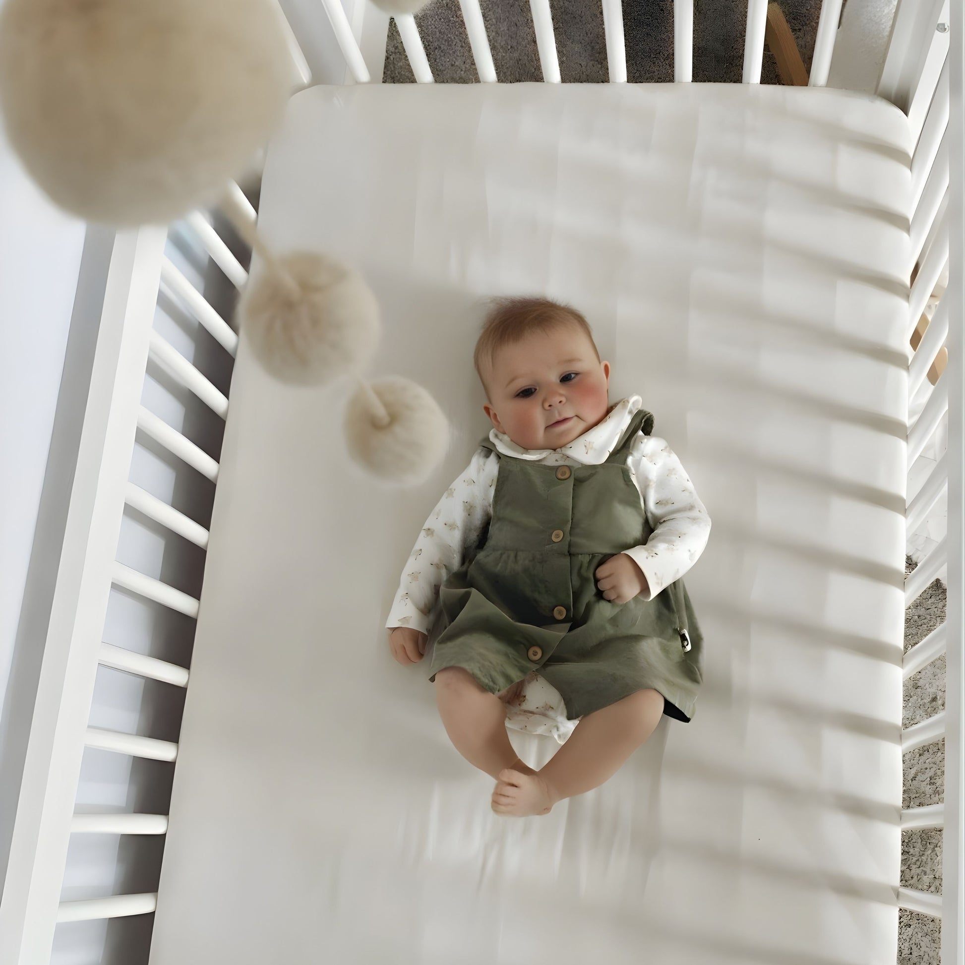 A baby in a green dress and white shirt lies on their back in the dadada baby Soho Convertible Crib, gazing at pom-pom mobile toys above. The nursery features cozy elements and subtle mid-century style accents.
