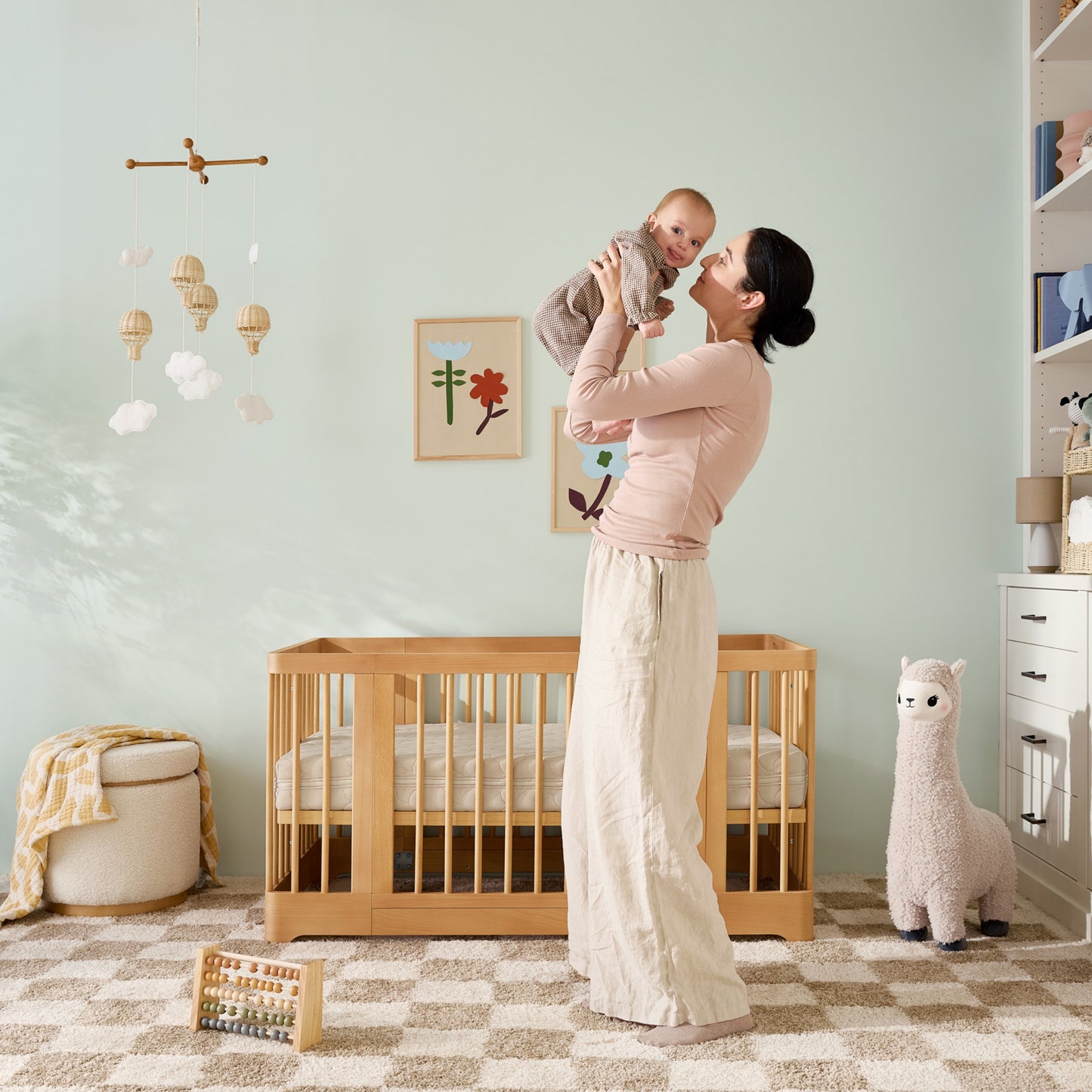 A woman stands in a nursery holding a smiling baby. Behind her is the Newton Baby Newton Nest Convertible Crib (GREENGUARD Gold Certified), wall art, a plush llama, and a mobile. The softly colored room has checkered carpet and children’s toys.