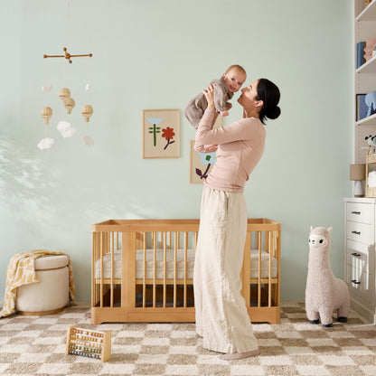 A woman stands in a nursery holding a smiling baby. Behind her is the Newton Baby Newton Nest Convertible Crib (GREENGUARD Gold Certified), wall art, a plush llama, and a mobile. The softly colored room has checkered carpet and children’s toys.