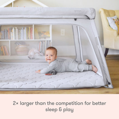 A smiling baby lies on their stomach in the Newton Baby Deluxe Travel Crib & Play Yard, featuring mesh sides and a breathable mattress. Text below reads: 2× larger than competitors for better sleep & play.