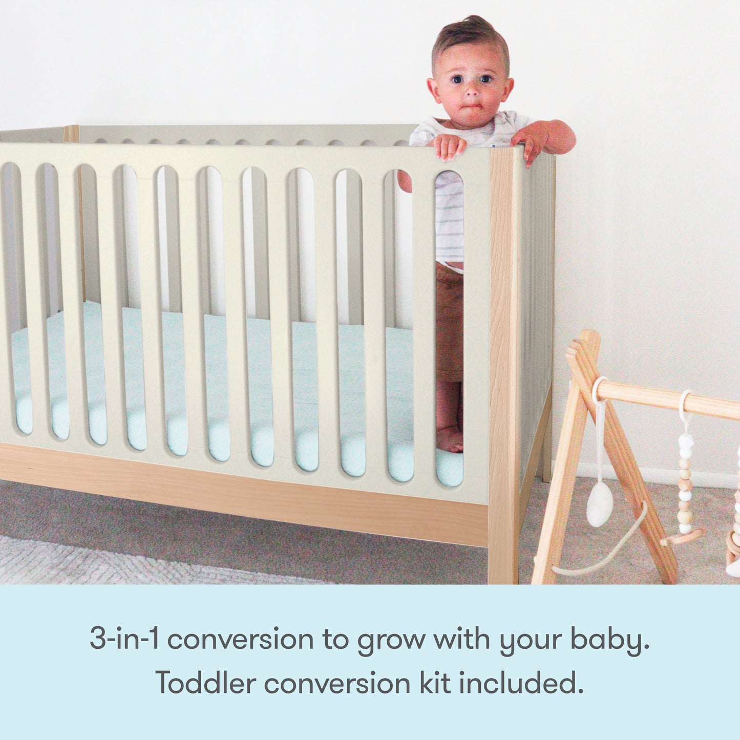 A baby stands in the Newton Galileo Convertible Crib by Newton Baby with a pale blue mattress in a minimal nursery. A wooden baby gym sits nearby. Text below reads: 3-in-1 convertible crib grows with your baby; toddler bed kit included.