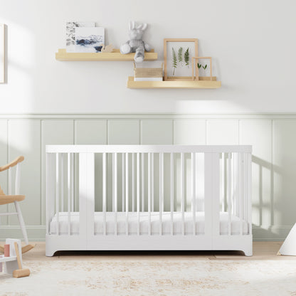 A GREENGUARD Gold certified, white Newton Baby Newton Nest Convertible Crib with a mattress stands against a pastel green and white wall. Above it, two wooden shelves display framed art, books, a teddy bear, and decor. A rocking chair is nearby on a light rug.