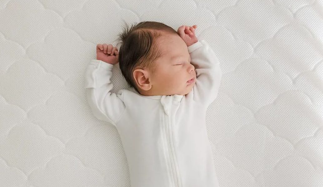 High-angle close-up view of a newborn baby lying on a white crib mattress. The baby is wearing a white, long-sleeved, zippered onesie. The baby's head and arms are turned to one side, and the baby appears to be sleeping soundly. The crib mattress has a textured, quilted pattern in a light, off-white color.