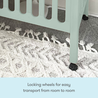 A close-up of a Newton Baby Galileo Mini Crib with Newton Mattress leg shows the black locking wheel on a textured white rug, emphasizing the mobility feature. Text: Locking wheels let you easily move the crib from room to room.