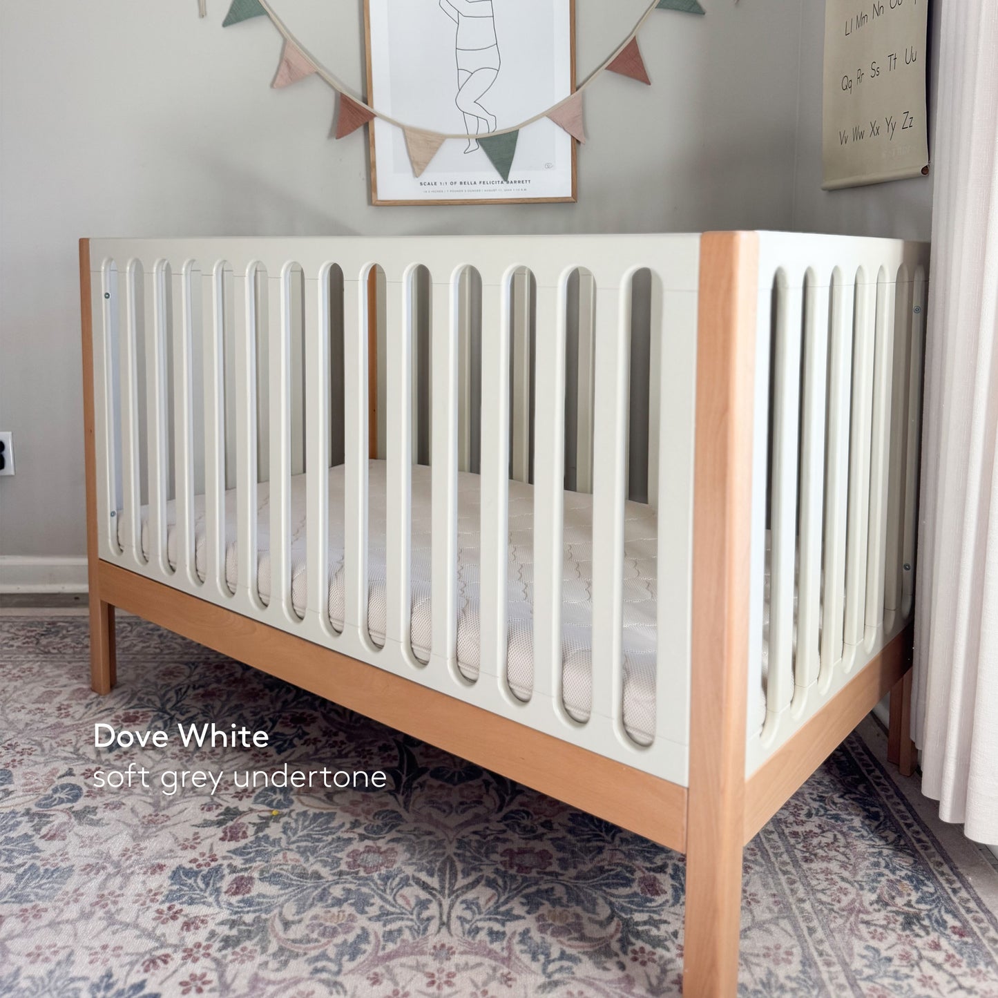 The Newton Baby Newton Galileo Convertible Crib, featuring white slats and natural beech wood legs, sits on a patterned rug in a softly lit nursery. Text on the image reads: Dove White, soft grey undertone.