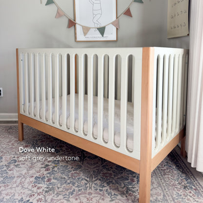 The Newton Baby Newton Galileo Convertible Crib, featuring white slats and natural beech wood legs, sits on a patterned rug in a softly lit nursery. Text on the image reads: Dove White, soft grey undertone.