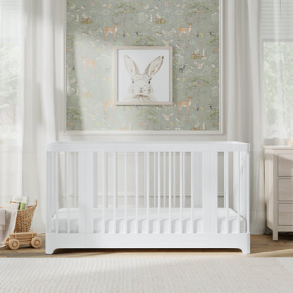 A white Newton Nest Convertible Crib by Newton Baby stands in a bright nursery with forest-themed wallpaper, a bunny picture above the crib, light curtains, and a wooden toy cart on the floor. This GREENGUARD Gold certified piece ensures both safety and style for your little one’s space.