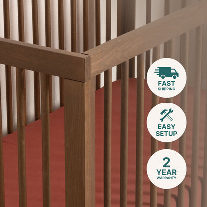 Close-up of the dadada baby Soho Convertible Crib, a mid-century style 3-in-1 crib with vertical slats and a rust-colored mattress. Icons highlight fast shipping, easy setup, and a 2-year warranty.