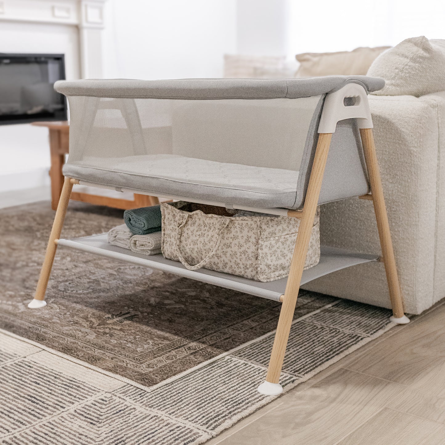 essential bassinet next to a couch in a living room, storage space below bassinet filled with sheets and other baby care essentials. 999-8402-GRY