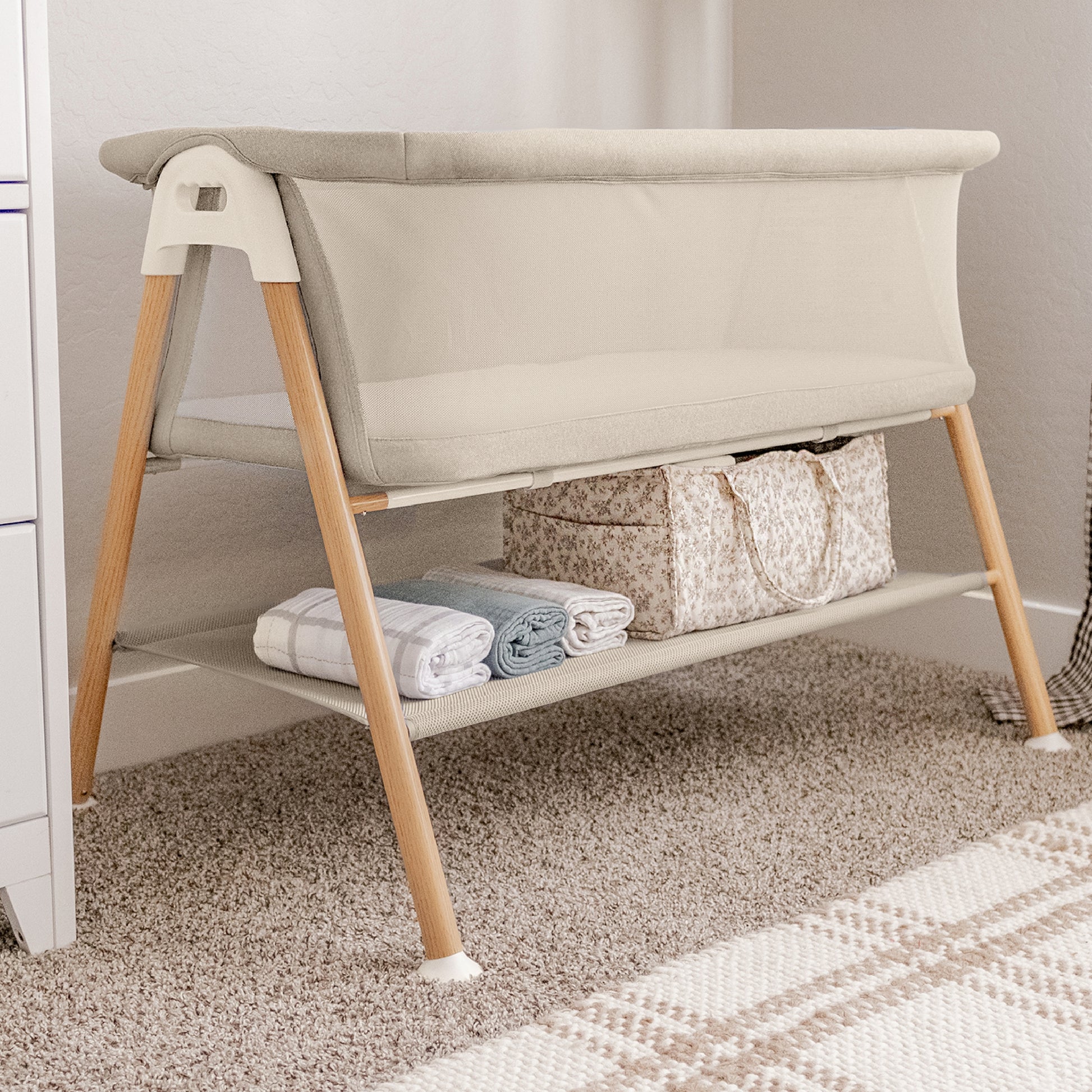 The Newton Baby Essential Bassinet in beige, featuring wooden legs, is placed on a carpeted floor. The bottom shelf stores a patterned bag, rolled blankets, and a folded cloth. A plaid rug is partly visible in front. 999-8402-SAN