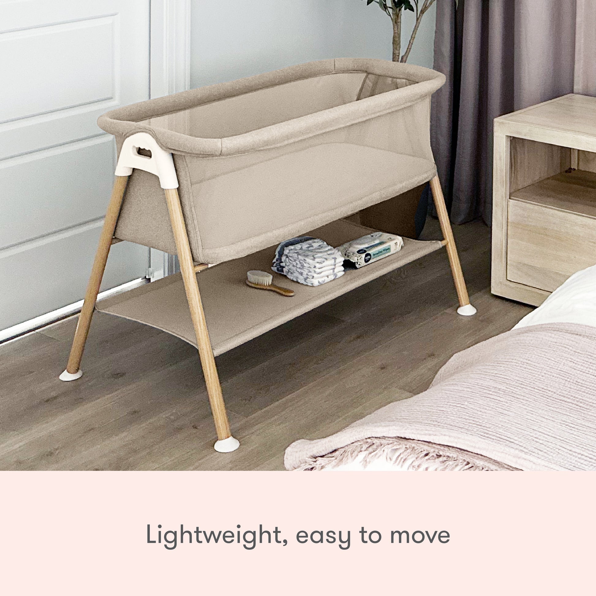 The Newton Baby Essential Bassinet, featuring breathable mesh sides and wooden legs in beige, sits beside a bed. Lightweight and easy to move, it provides storage for essentials like diapers, wipes, and a brush underneath.