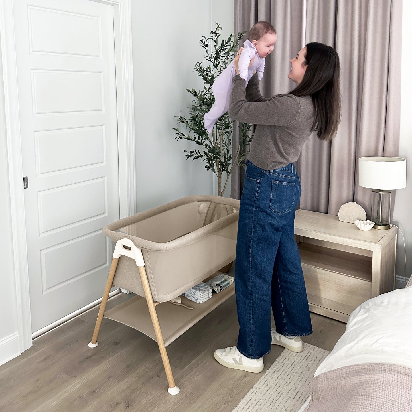 A woman smiles as she lifts a baby in a purple onesie above the Newton Baby Essential Bassinet, which features breathable mesh sides. The bassinet is in a bedroom with light walls, a wooden nightstand, lamp, and potted plant.