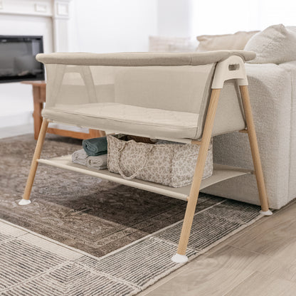 The Newton Baby Essential Bassinet with wooden legs stands by a cream sofa. Its breathable mesh sides and storage shelf hold rolled blankets and a patterned bag, adding function and comfort to this cozy living room.