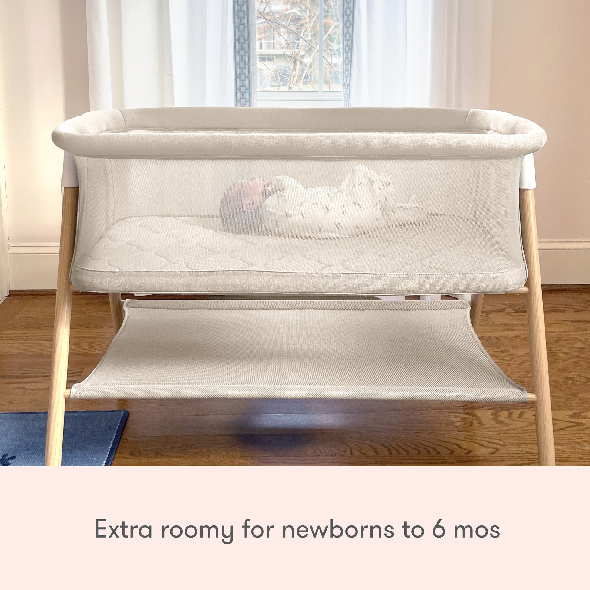 A baby rests in the Newton Baby Essential Bassinet, a spacious, non-toxic sleeper with light wood legs by a sunlit window. Text below reads: Extra roomy for newborns to 6 months.