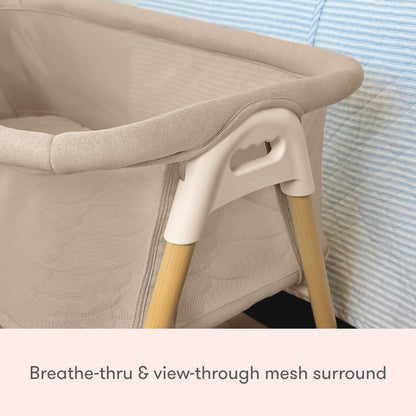 A close-up of the Newton Baby Essential Bassinet, featuring breathable, view-through mesh sides and wooden legs, is placed next to a blue-striped surface.