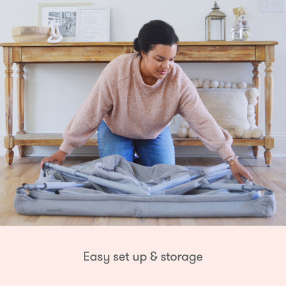 A woman kneels on a wooden floor, folding the Newton Baby Deluxe Travel Crib & Play Yard in front of a wooden console table with decor. A cushion is underneath. Text below reads: Easy setup & storage with breathable mattress.