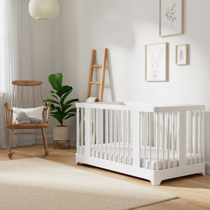 Bright nursery room featuring the Newton Baby Newton Nest Convertible Crib, which is GREENGUARD Gold certified. Room also includes a wooden chair topped with a cat pillow, a potted plant, ladder shelf, and bunny wall art. Light wood floors and neutral décor create a calm, cozy atmosphere.