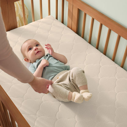 An adult adjusts a baby’s clothes as the baby lies on a cream-colored mattress in the Newton Baby Newton Nest Convertible Crib, which features GREENGUARD Gold certification. The baby wears a blue shirt and beige pants.