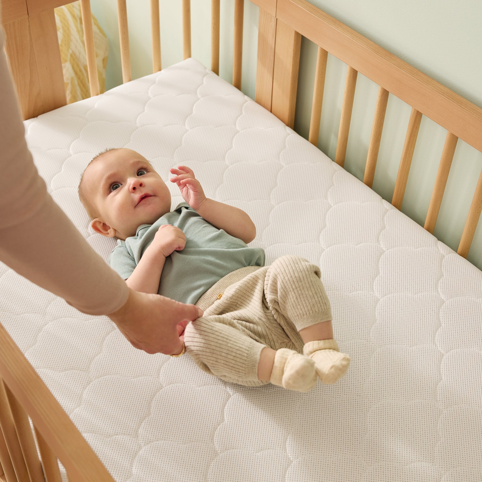 A baby in a light blue top and beige pants lies on their back in a Newton Nest Convertible Crib by Newton Baby, featuring a quilted white mattress, as an adult hand gently holds the baby's feet.