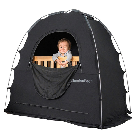 Baby in crib peeking out through window into slumberpod tent, big black tent over wooden crib with baby inside. 
