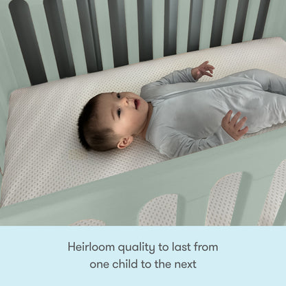 A baby in a light gray onesie lies on a white Newton Mattress inside the Newton Galileo Mini Crib by Newton Baby. Text below reads, Heirloom quality to last from one child to the next.