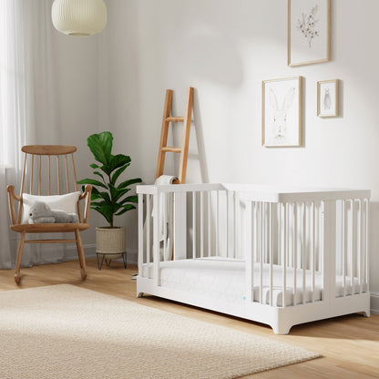 Sunlight streams into a bright nursery featuring the Newton Nest Convertible Crib by Newton Baby, which is GREENGUARD Gold certified and finished in white. The room also includes a wooden rocking chair holding a stuffed toy, a ladder shelf, potted plant, beige rug, and animal-themed wall art adorning light-colored walls. White curtains frame the window, letting in plenty of natural light.