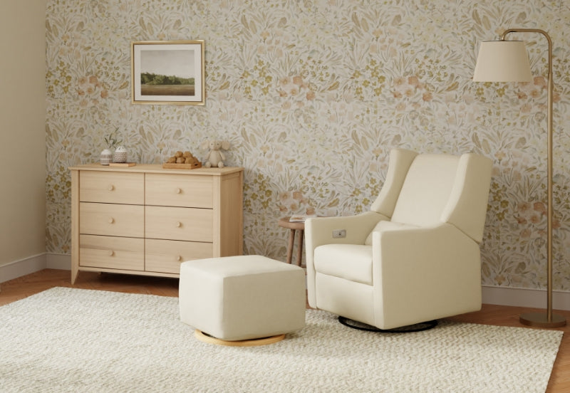 Elegant nursery featuring a beige glider chair with matching ottoman, wooden dresser, floral wallpaper, and soft lighting.