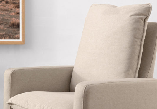 Beige nursery rocking chair with a comfortable back cushion, positioned against a wall near a framed picture.