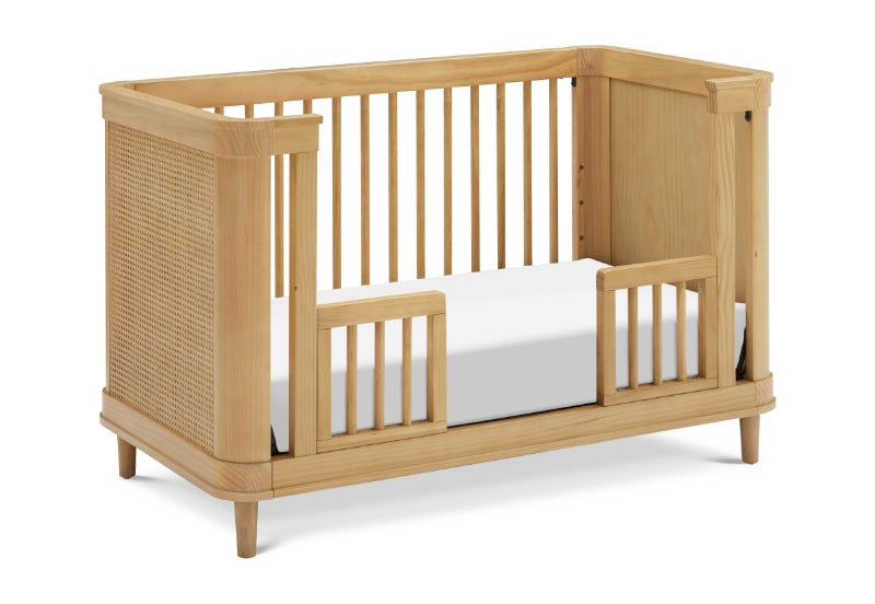 A natural wood baby crib featuring a slatted design on three sides and a woven rattan panel on one end, shown with a white mattress.