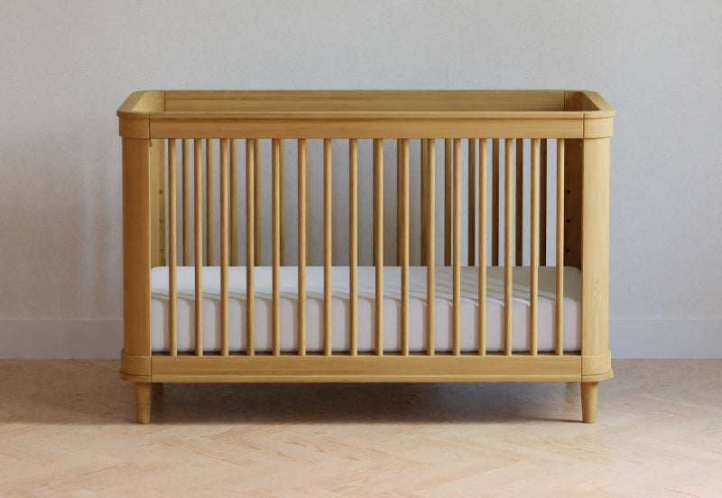 A simple wooden baby crib with a white mattress, placed against a neutral wall and light wood floor. 999-8607-HNY