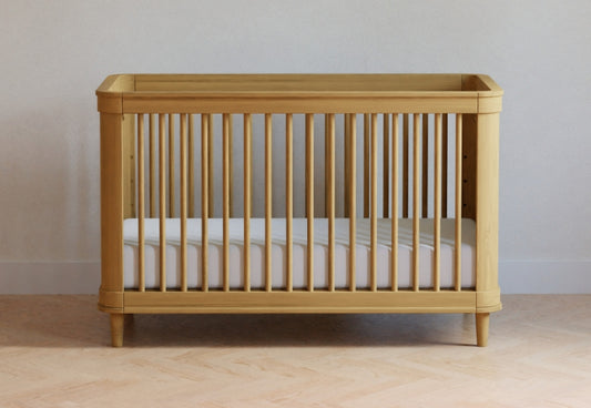 A simple wooden baby crib with a white mattress, placed against a neutral wall and light wood floor. 999-8607-HNY