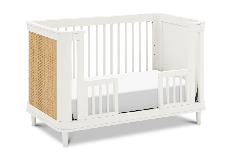 A modern white baby crib with a wooden slat design on one side.