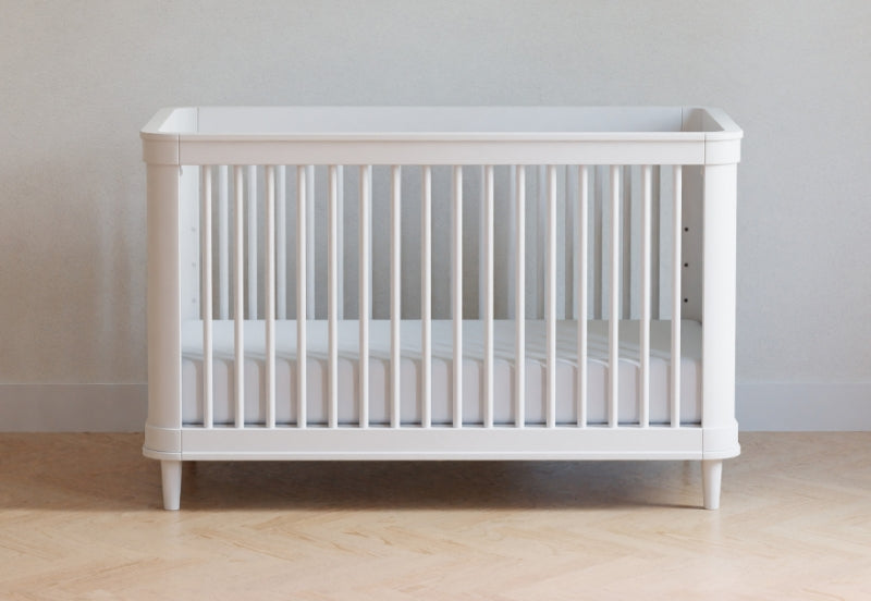 White baby crib against a neutral wall on a wooden floor. 999-8607-WHT