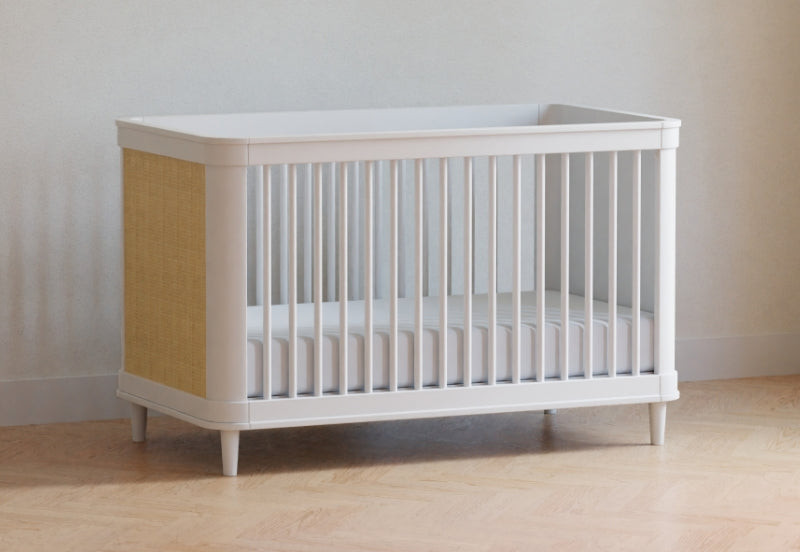 White modern crib with beige accents positioned against a neutral wall on a wooden floor, no text visible.
