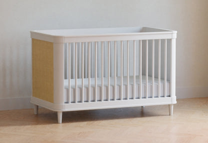 White modern crib with beige accents positioned against a neutral wall on a wooden floor, no text visible.