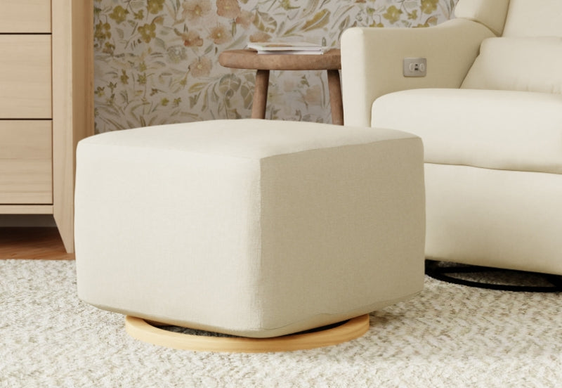 Beige nursery glider ottoman on a wooden base, in a cozy room with floral wallpaper and elegant furniture.