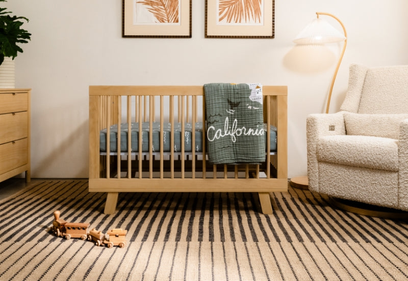 Cozy nursery room with a wooden crib adorned with a "California" themed blanket, a playful toy airplane on the floor, and a comfortable armchair beside a warm lamp.