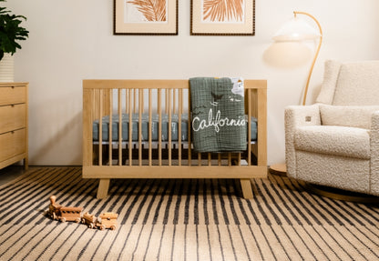 Cozy nursery room with a wooden crib adorned with a "California" themed blanket, a playful toy airplane on the floor, and a comfortable armchair beside a warm lamp.