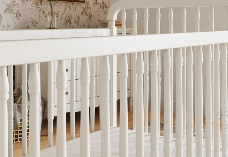 Close-up view of a white baby crib with elegant spindle details in a nursery with vintage-style wallpaper.