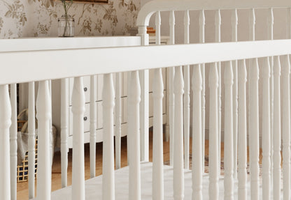 Close-up view of a white baby crib with elegant spindle details in a nursery with vintage-style wallpaper.