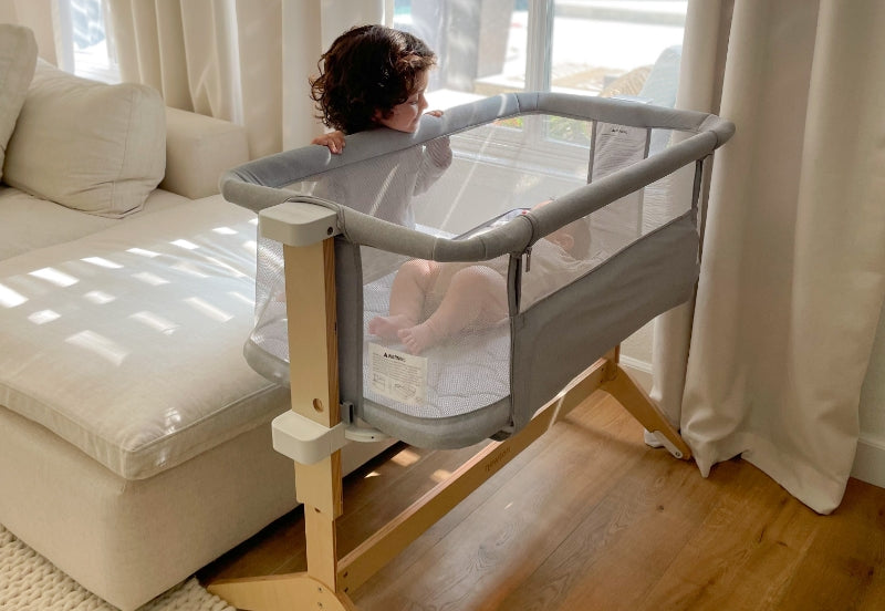 A toddler playfully standing in a modern gray baby crib next to a sunlit window, looking out through white curtains.