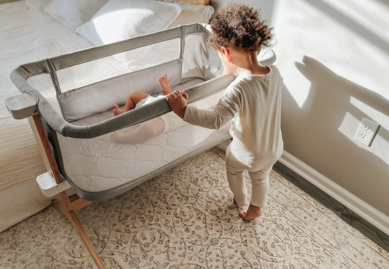 Toddler reaching out to a baby resting in a modern bedside bassinet in a sunlit room.