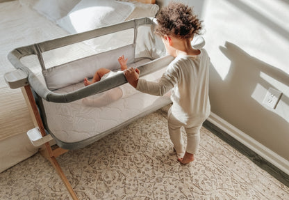Toddler reaching out to a baby resting in a modern bedside bassinet in a sunlit room.