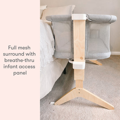 A modern gray baby bassinet with wooden legs placed in a serene room, featuring the text 'full mesh surround with breathe-thru infant access panel."
