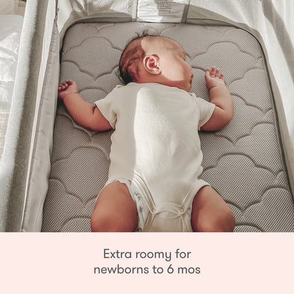 A baby sleeps on its back in the spacious Newton Baby Bassinet & Bedside Sleeper with a cloud-patterned Wovenaire mattress. Text below reads: “Extra roomy for newborns to 6 mos.”.