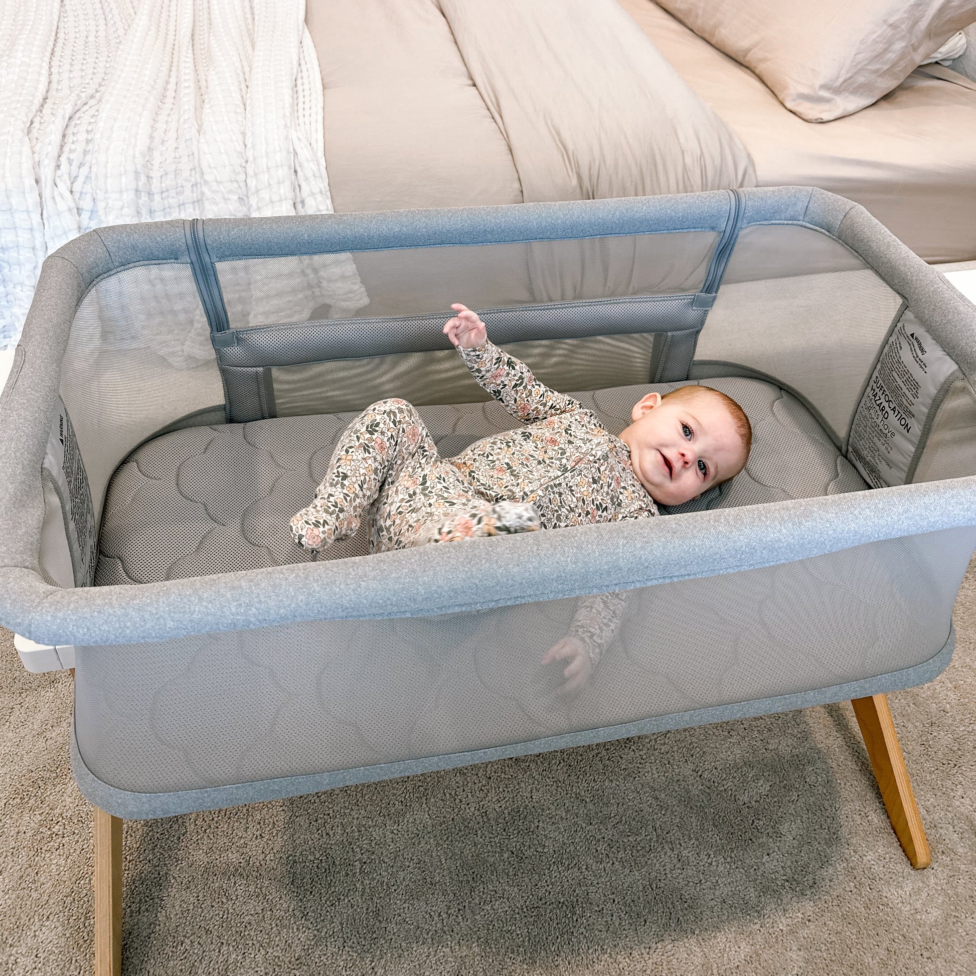 A baby in a floral onesie smiles in the Newton Baby Bundle 3 BFCM bassinet, which features wooden legs and gray mesh sides, set beside a neatly made bed in a cozy bedroom.