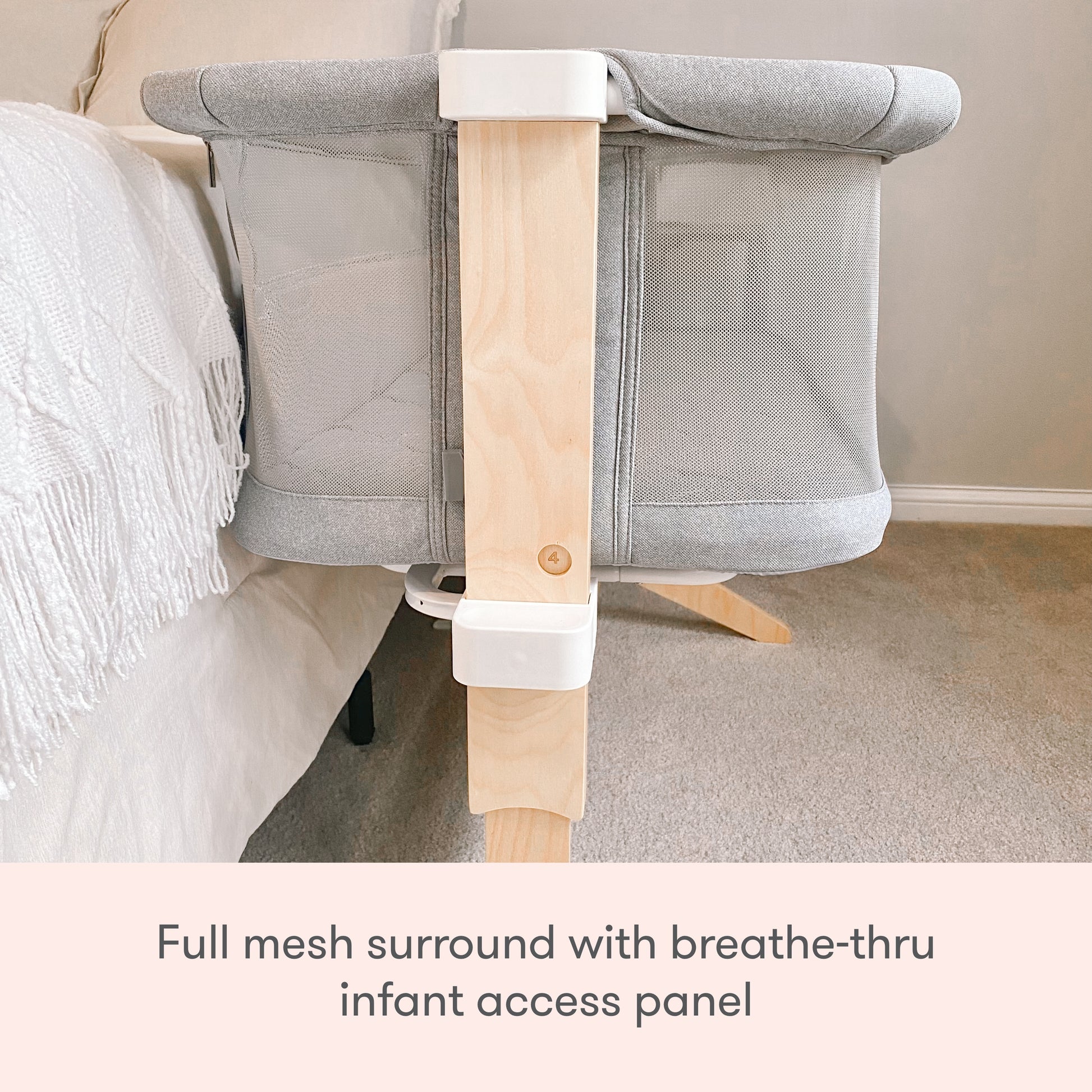 A Newton Baby Bassinet & Bedside Sleeper sits next to a bed, featuring a grey, breathable full mesh surround with wooden support and a breathe-thru infant access panel. Text: Full mesh surround with breathe-thru infant access panel.