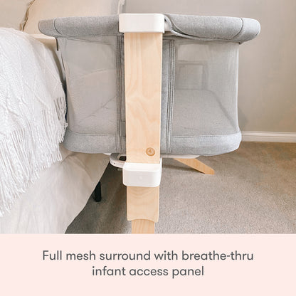 A Newton Baby Bassinet & Bedside Sleeper sits next to a bed, featuring a grey, breathable full mesh surround with wooden support and a breathe-thru infant access panel. Text: Full mesh surround with breathe-thru infant access panel.