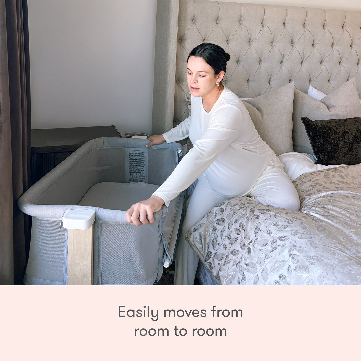 A woman in white pajamas sits on a bed and adjusts the Newton Baby Bassinet & Bedside Sleeper in a cozy, well-lit bedroom. Text below reads, Easily moves from room to room.