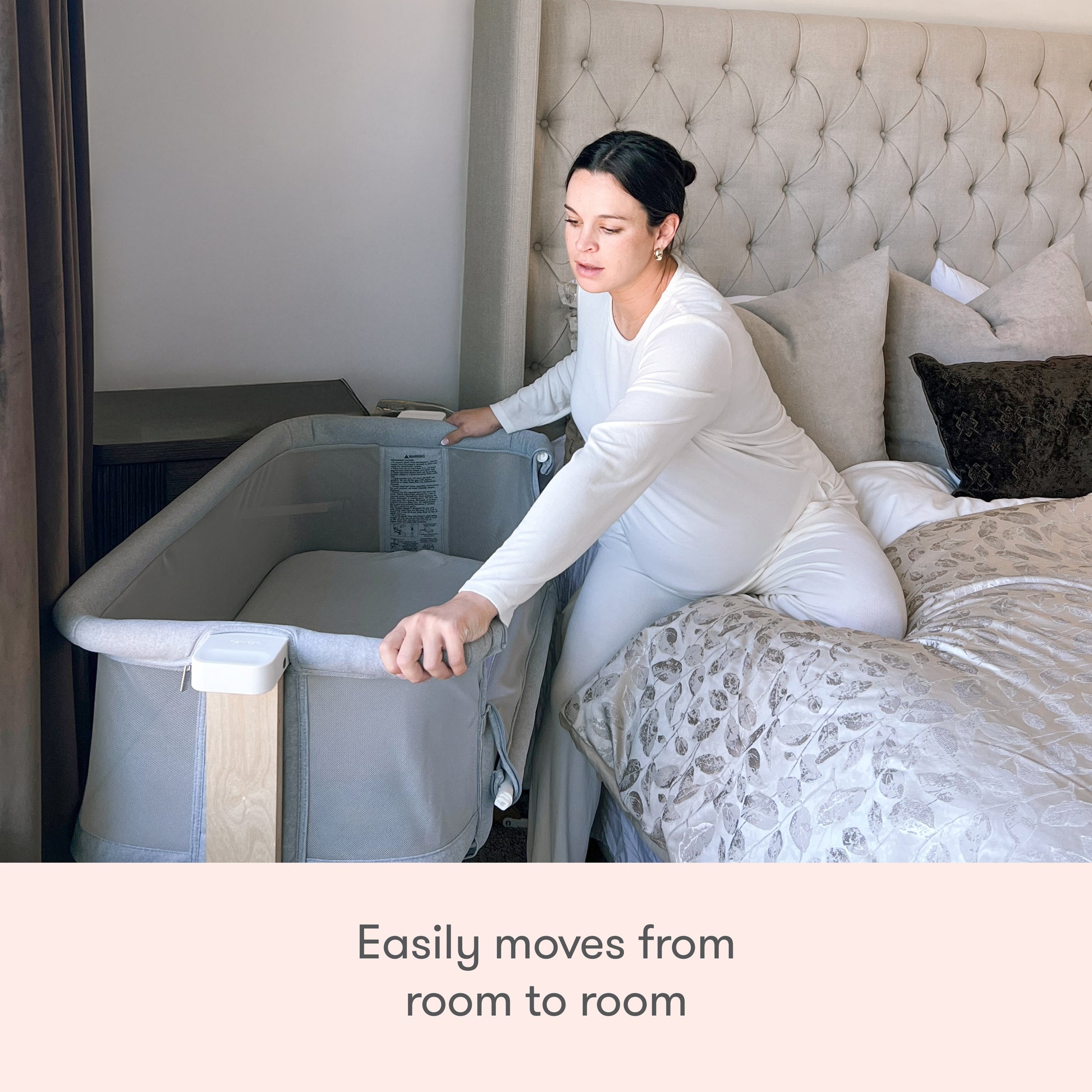A woman in white pajamas sits on a bed and adjusts the Newton Baby Bassinet & Bedside Sleeper in a cozy, well-lit bedroom. Text below reads, Easily moves from room to room.